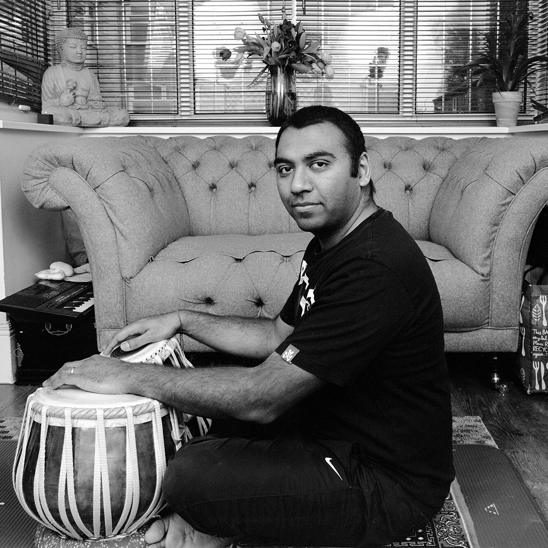 A cross-legged tabla player sits side-on to us staring out of the frame. His tabla are in front of him and his hands are on them