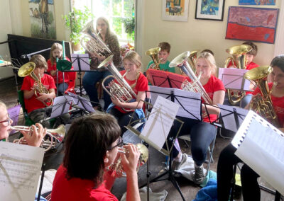 A group of young people of different ages are playing brass instruments in a circle in a bright sunny room. There are music stands and music in front of each one.