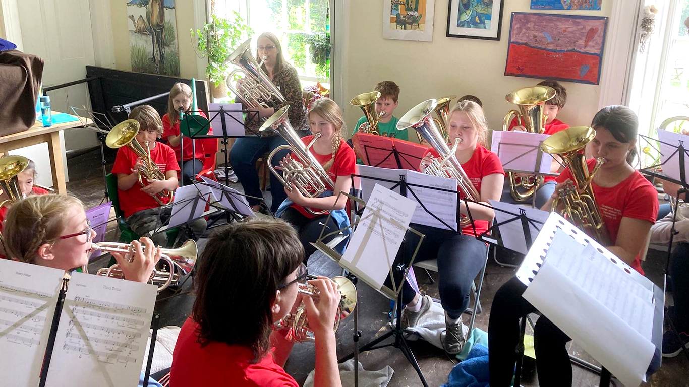 A group of young people of different ages are playing brass instruments in a circle in a bright sunny room. There are music stands and music in front of each one.