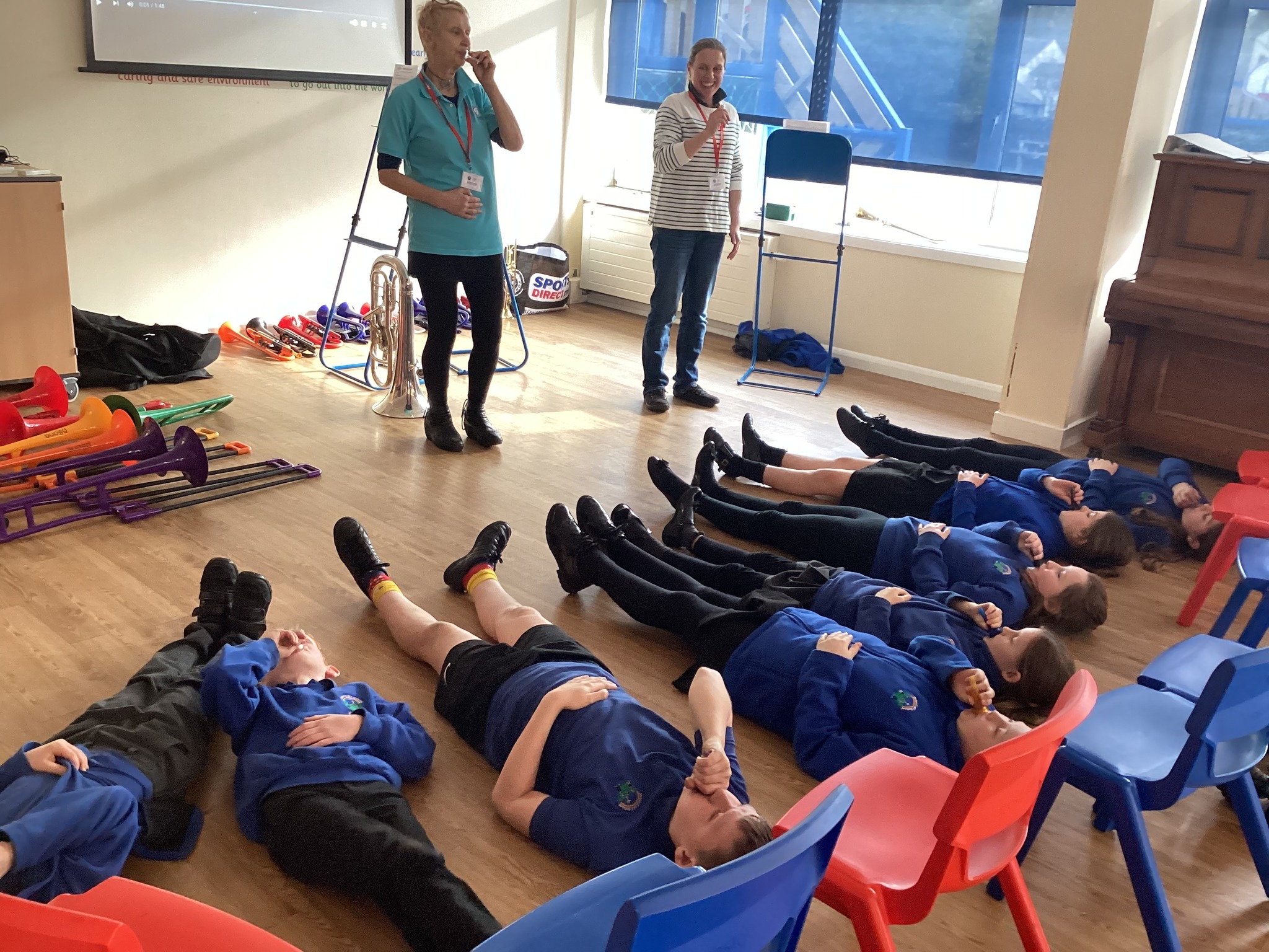 Young people lie on the floor holding brass mouthpieces as music leader explains how to blow effectively