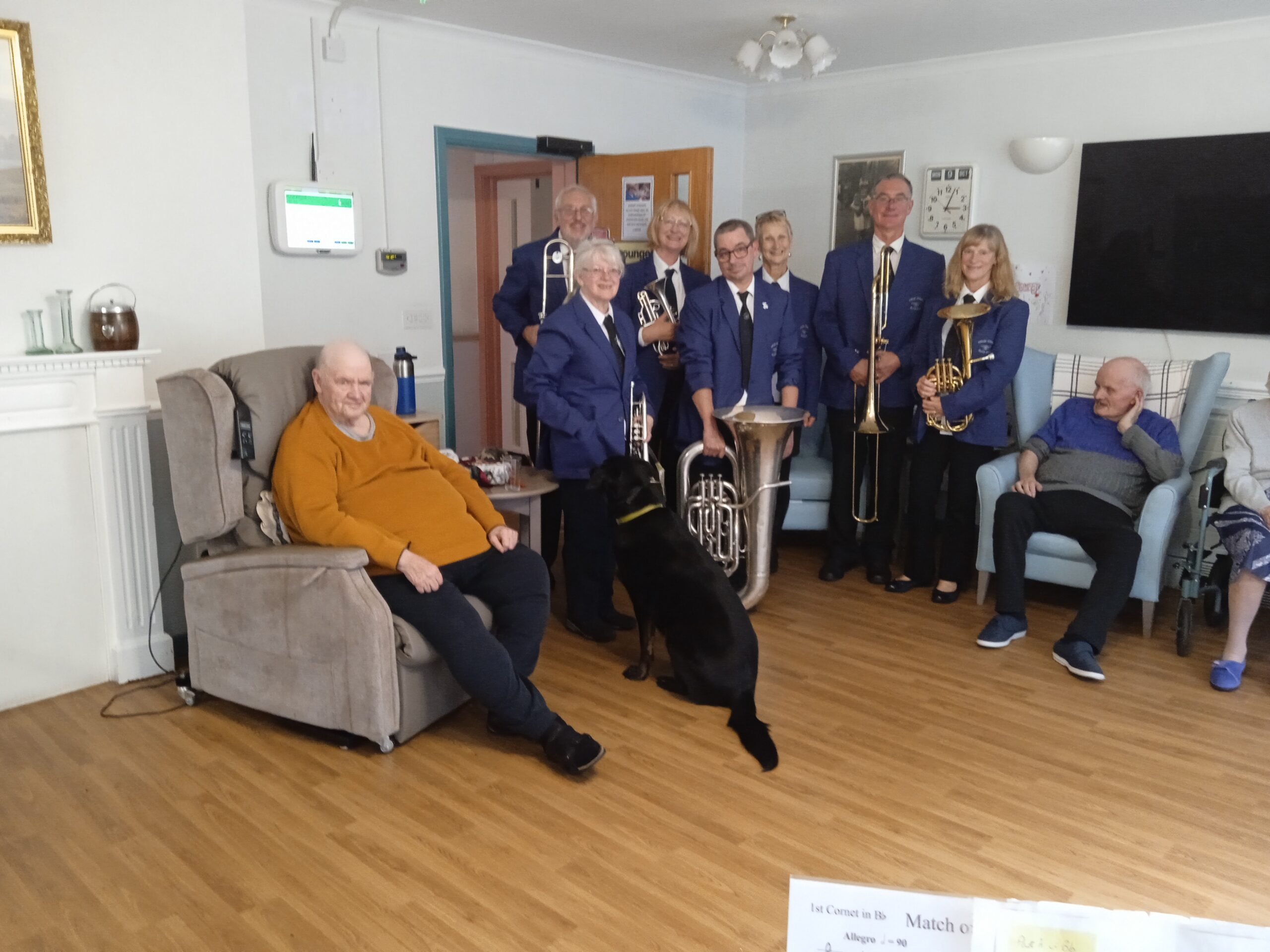 A group of elderly people sit in comfy chairs while band players stand behinf them, wearing smart blue uniforms and smiling