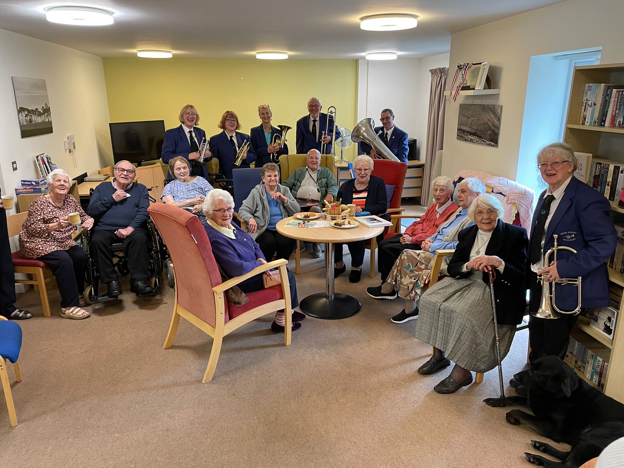 A group of elderly people sit in comfy chairs while band players stand behinf them, wearing smart blue uniforms and smiling